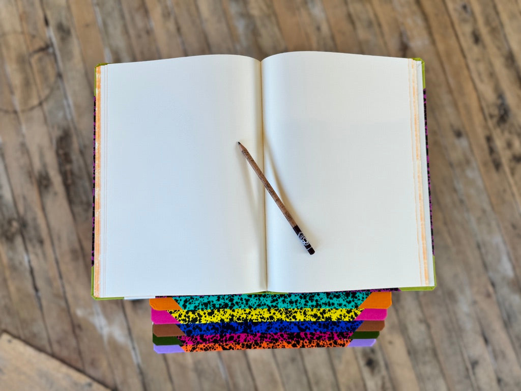 An open blank notebook with white pages lies on a rustic wooden surface, with a pencil resting across the center and a stack of colorful patterned notebooks or book covers visible underneath.