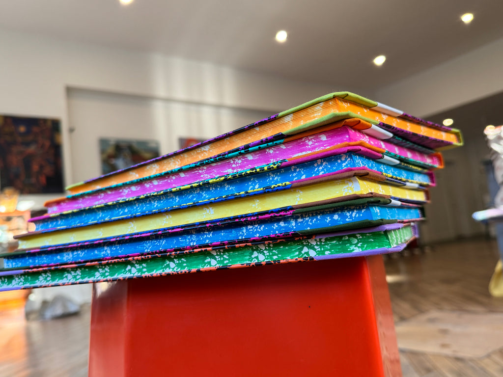 A stack of colorful patterned notebooks or art books displayed on a red pedestal in an indoor gallery or studio space.