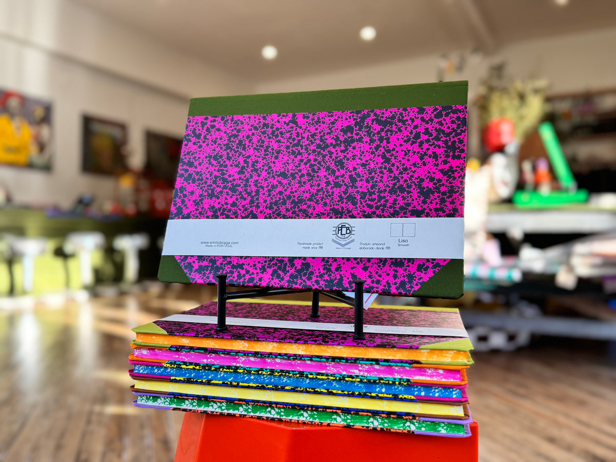 A stack of colorful composition notebooks displayed on a small stand in a blurred indoor store or office setting. The top notebook features a bright pink and black speckled cover with green borders, and a white paper band is wrapped around the middle.