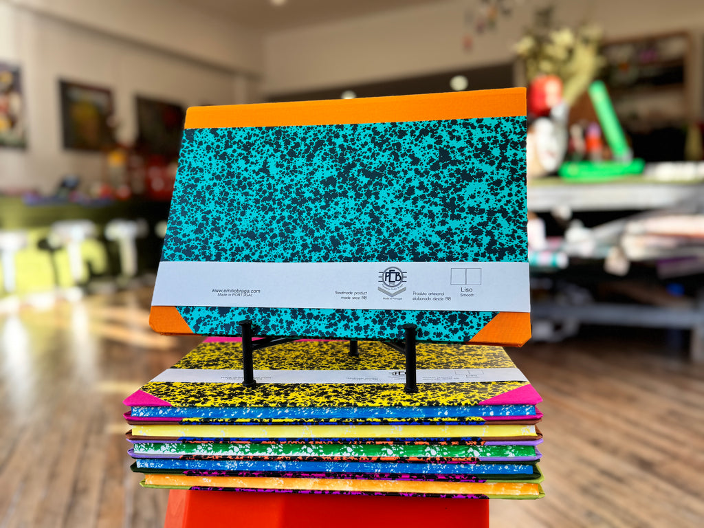 A stack of colorful composition notebooks displayed on small black stands in a blurred indoor shop or office environment. The top notebook features a turquoise and black speckled cover with orange binding, and a white paper band around the middle shows the 'DFP' brand logo.