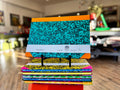 A stack of colorful composition notebooks displayed on small black stands in a blurred indoor shop or office environment. The top notebook features a turquoise and black speckled cover with orange binding, and a white paper band around the middle shows the 'DFP' brand logo.