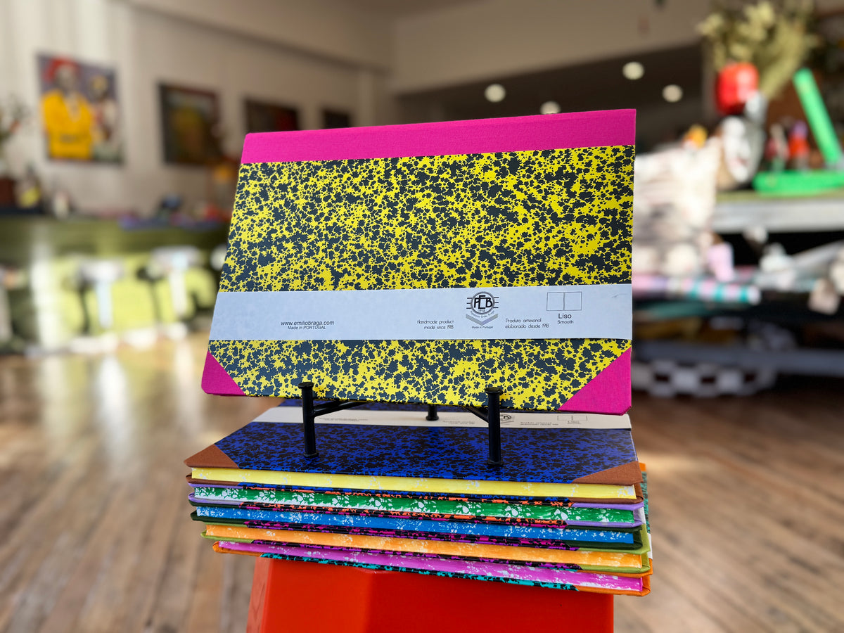 A stack of colorful composition notebooks with yellow, black, blue, and green covers displayed on a small stand in a blurred indoor store or office setting.