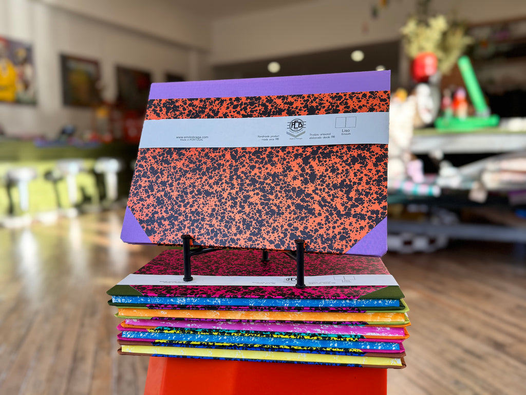 A stack of colorful composition notebooks displayed on a small orange stand in a blurred indoor shop or classroom setting. The top notebook features a purple and black speckled cover with a white paper band around the middle.