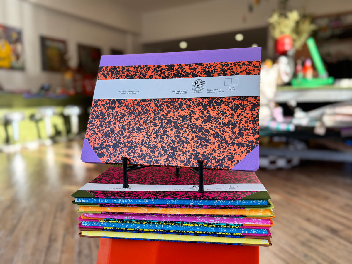 A stack of colorful composition notebooks displayed on a small orange stand in a blurred indoor shop or classroom setting. The top notebook features a purple and black speckled cover with a white paper band around the middle.