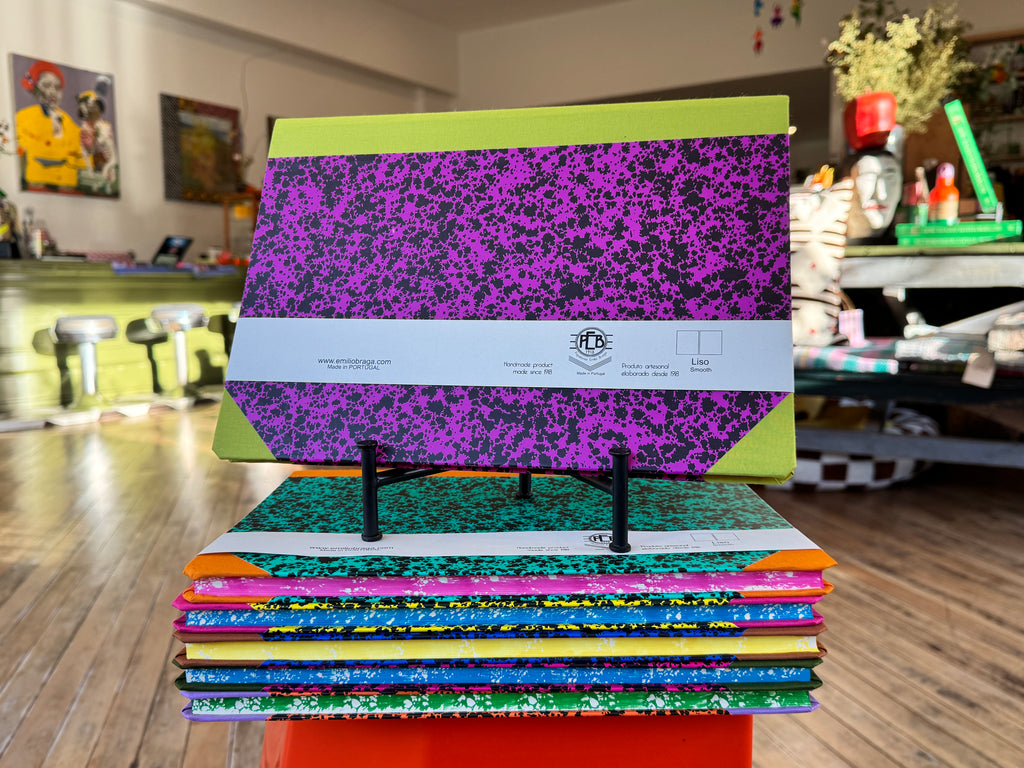 A stack of colorful composition notebooks displayed on a small stand in a retail store. The top notebook has a bright lime green cover with a large purple and black speckled pattern, and a white paper band around the middle.