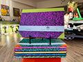 A stack of colorful composition notebooks displayed on a small stand in a retail store. The top notebook has a bright lime green cover with a large purple and black speckled pattern, and a white paper band around the middle.