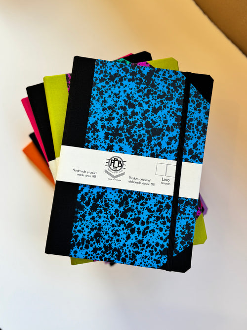 A stack of hardcover notebooks with black elastic closure bands and a white paper band around the top notebook. The top notebook has a bright blue and black splatter pattern, and the notebooks underneath show additional colors including lime green, pink, orange, and purple.