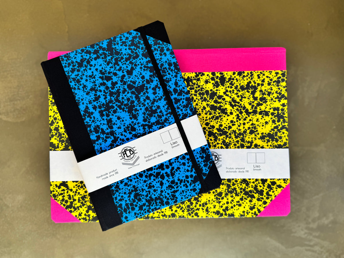 Two hardcover notebooks with speckled covers stacked on a textured beige surface. The top notebook has a blue and black speckled cover with a black elastic closure, and the bottom notebook has a yellow and black speckled cover with a hot pink corner accent.