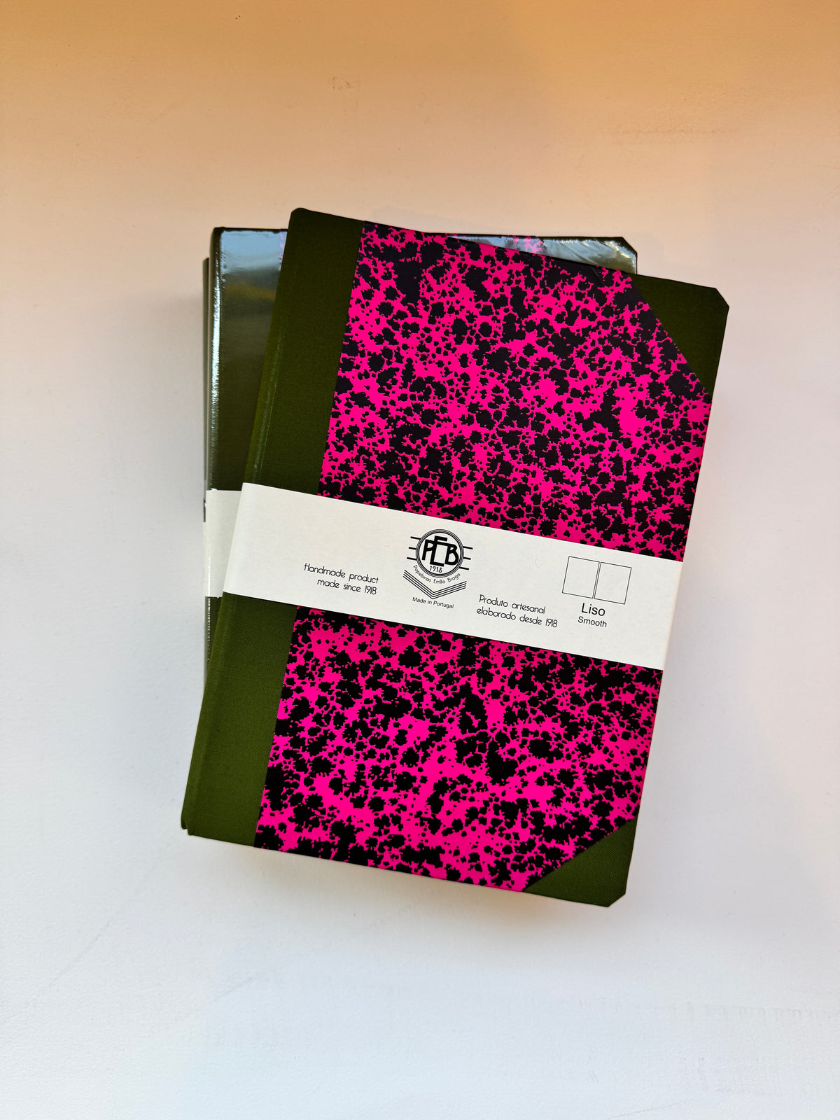 A stack of notebooks with a vibrant pink and black splatter pattern and dark green edges, wrapped in a white paper band with a visible 'The Paper Club' logo, on a plain white surface.