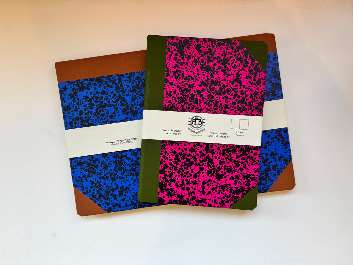 Two vintage-style composition notebooks with brown leather corners and white paper bands, displayed on a white surface. One notebook features a vibrant blue and black splatter pattern, and the other features a hot pink and black splatter pattern with an olive green spine.