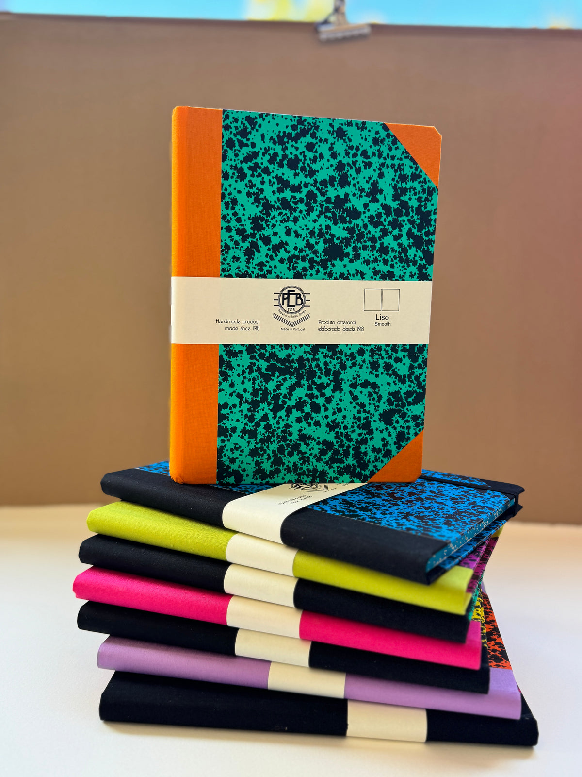 A stack of colorful notebooks with black-and-white marbled covers, orange spines, and cream paper bands, displayed on a tabletop against a tan background.