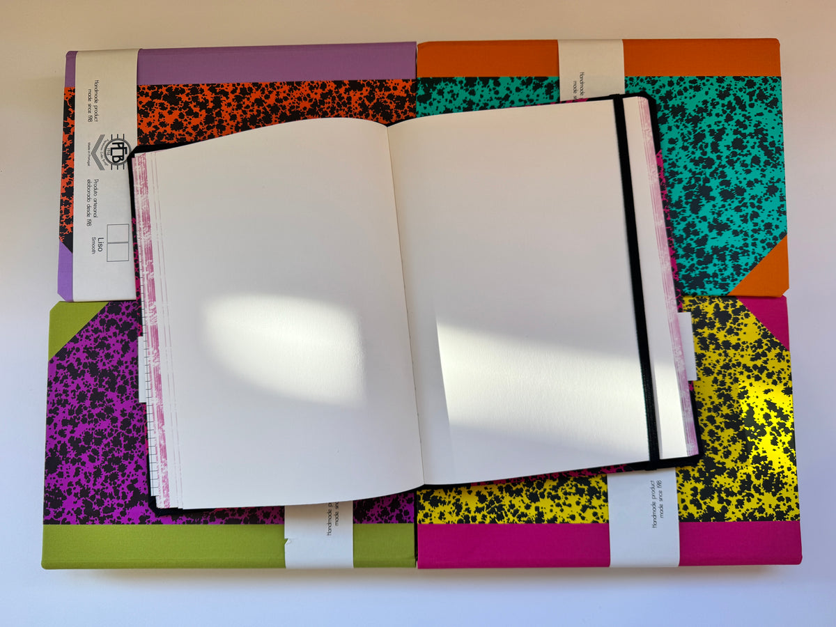 An open blank notebook with white pages lies on top of several closed notebooks with colorful speckled covers, photographed from above on a white background.