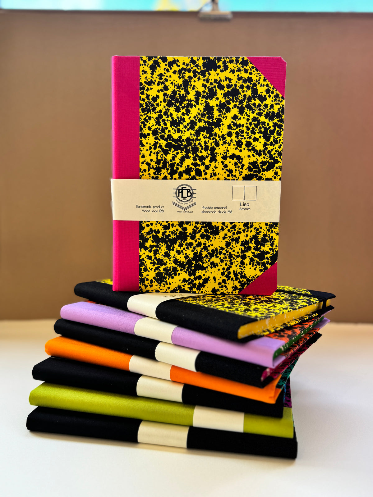 A stack of colorful notebooks with black and yellow speckled covers, some standing upright and others fanned out below, set against a plain brown background.