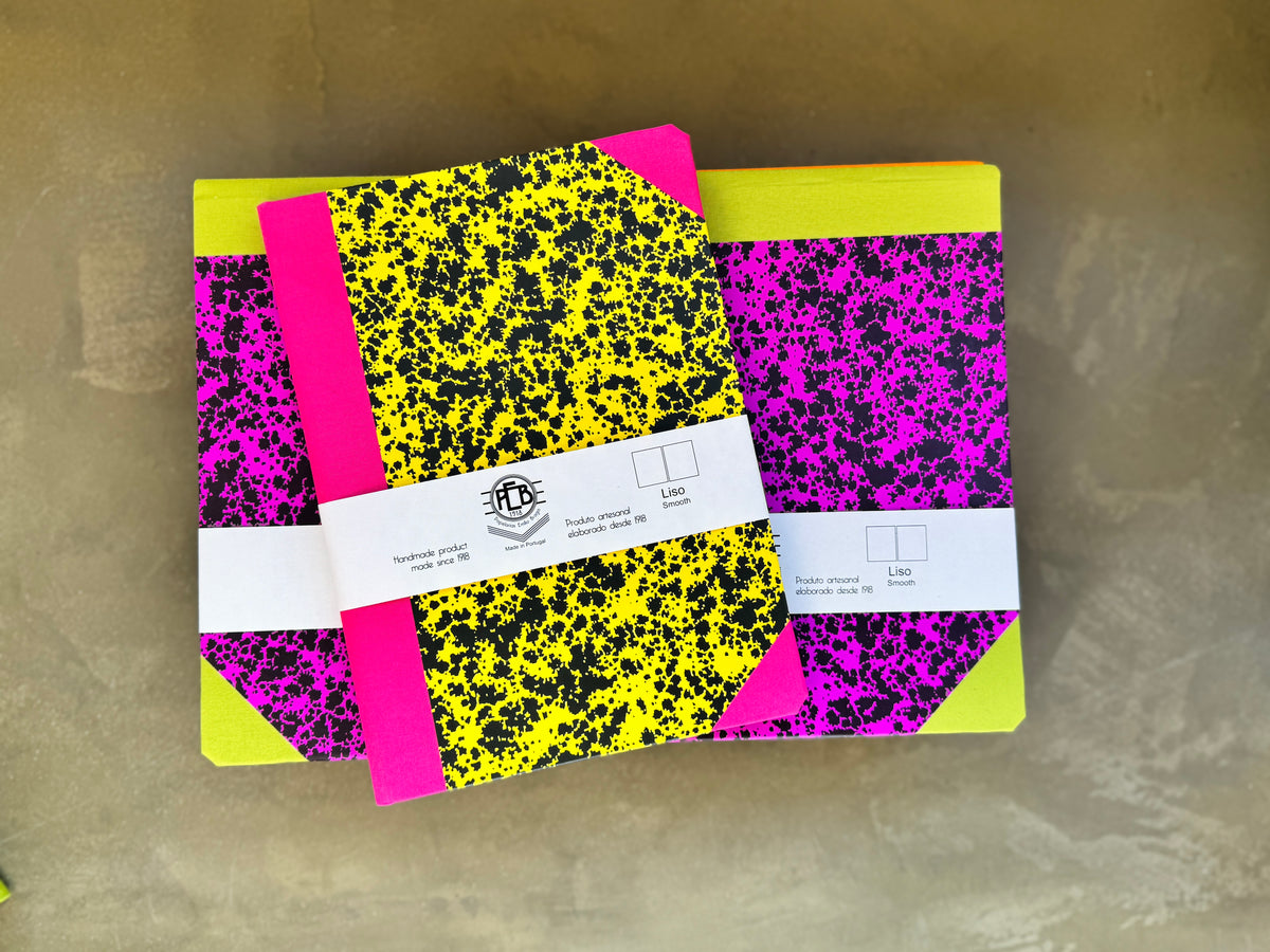 Two colorful composition notebooks with a black and neon yellow/slime green splatter pattern and hot pink binding, wrapped in a white paper band with a logo, on a textured gray surface.