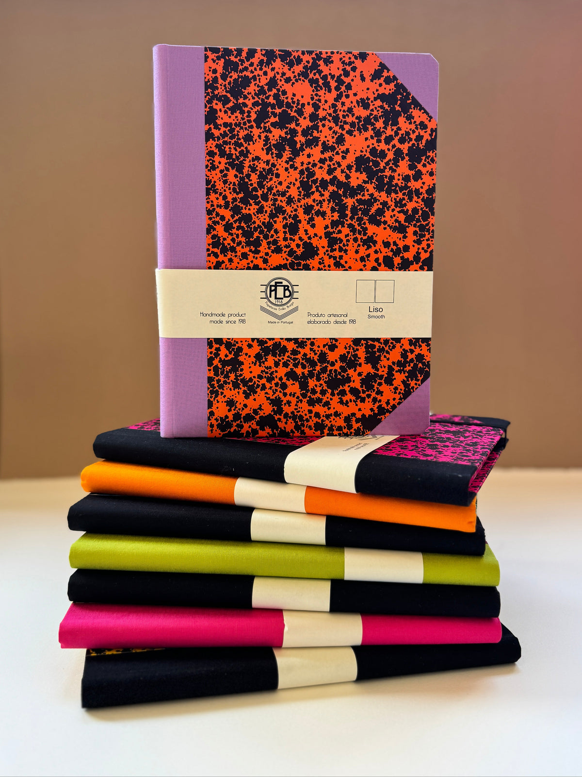 A stack of colorful notebooks with black and orange splatter patterns, purple, black, orange, lime green, pink, and black covers, wrapped in white paper bands with a circular logo in the center.