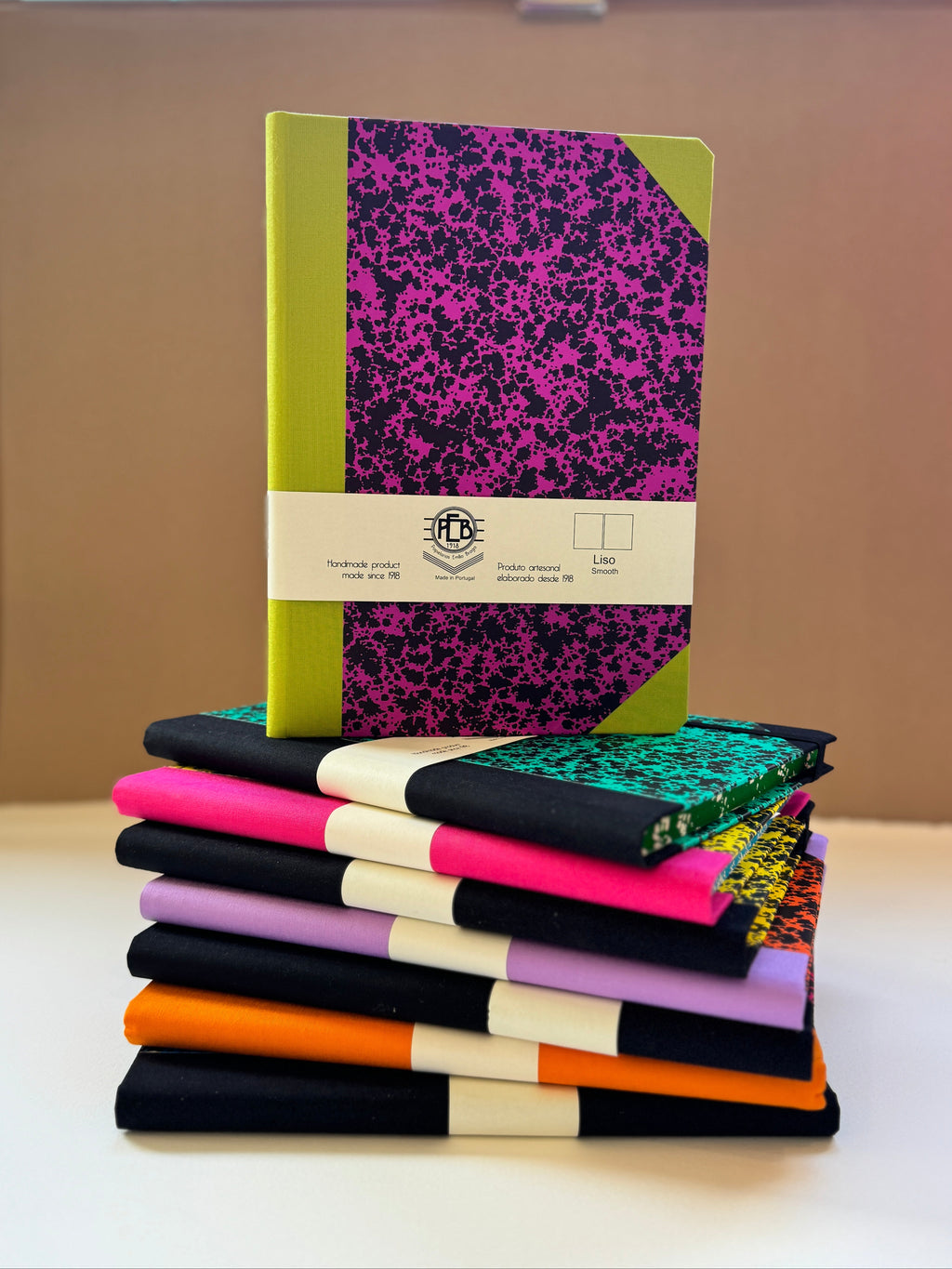 A stack of notebooks and journals displayed on a white surface against a beige background. The top notebook features a bright lime green cover with a black and magenta splatter pattern, wrapped in a white paper band.