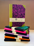 A stack of notebooks and journals displayed on a white surface against a beige background. The top notebook features a bright lime green cover with a black and magenta splatter pattern, wrapped in a white paper band.