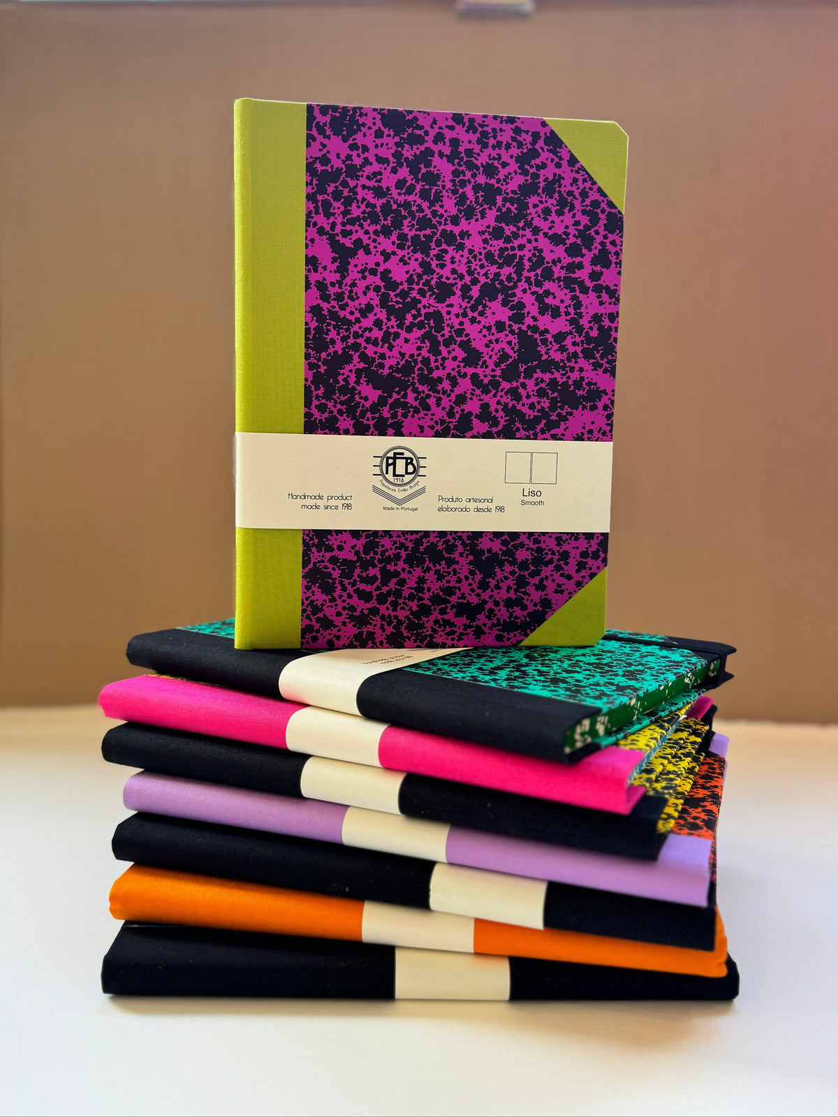 A stack of notebooks and journals displayed on a white surface against a beige background. The top notebook features a bright lime green cover with a black and magenta splatter pattern, wrapped in a white paper band.