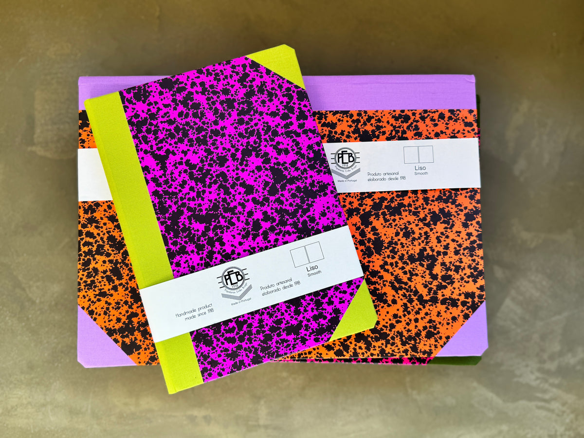 Two notebooks with vibrant orange, black, and purple speckled covers, stacked on top of each other on a textured gold surface. Each notebook has a lime green corner accent, a white paper band with a logo, and a purple edge.