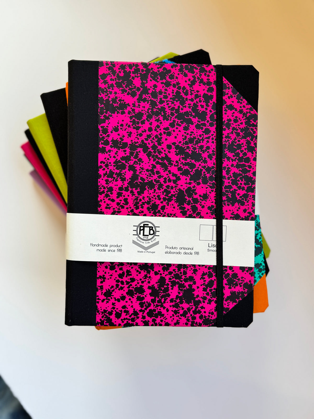 A stack of hardcover notebooks with a black and hot pink splatter pattern, wrapped in a white paper band with a visible FTD logo, set against a plain off-white background.