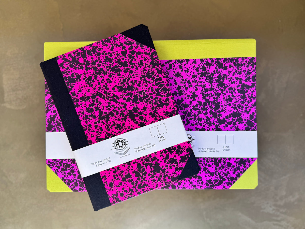 Two notebooks with a bright pink and black splatter pattern and yellow covers, wrapped in white paper bands with a logo, on a neutral background.