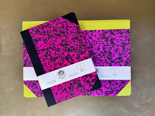 Two notebooks with a bright pink and black splatter pattern and yellow covers, wrapped in white paper bands with a logo, on a neutral background.