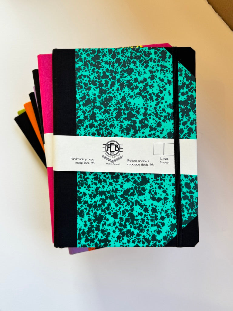A stack of notebooks with a black and turquoise splatter patterned cover, held together by a white paper band with the brand logo 'HTC'.