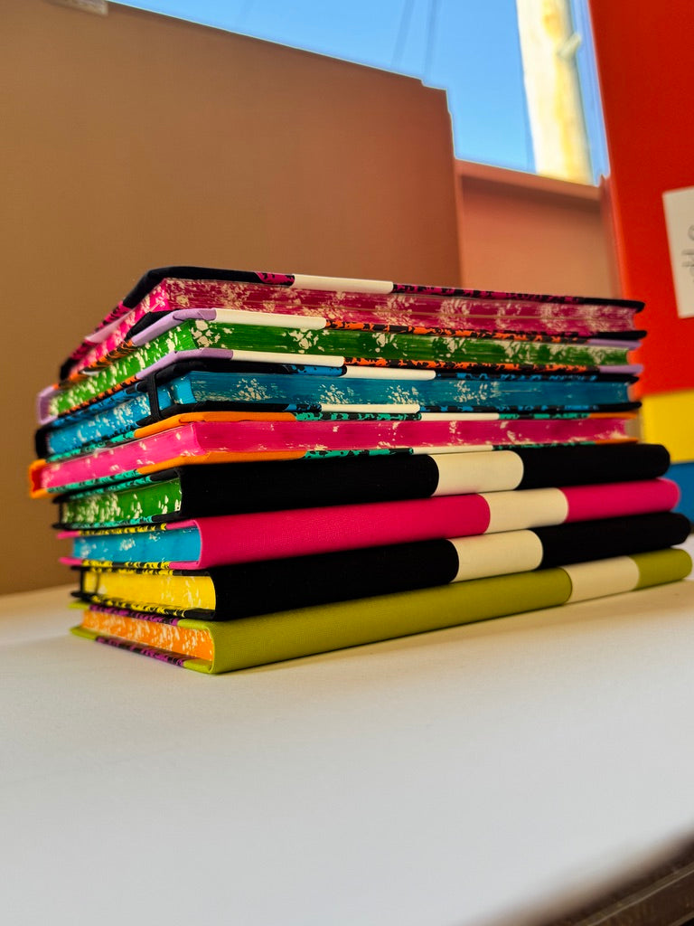 A stack of colorful patterned notebooks or journals with black, pink, green, blue, and yellow covers, arranged on a white surface.