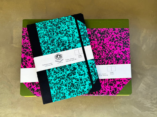 Two hardcover notebooks with black and neon pink marbled covers, stacked on top of each other on a gold-colored surface. The top notebook has a black elastic closure and a white paper band with the brand logo 'LMS'.