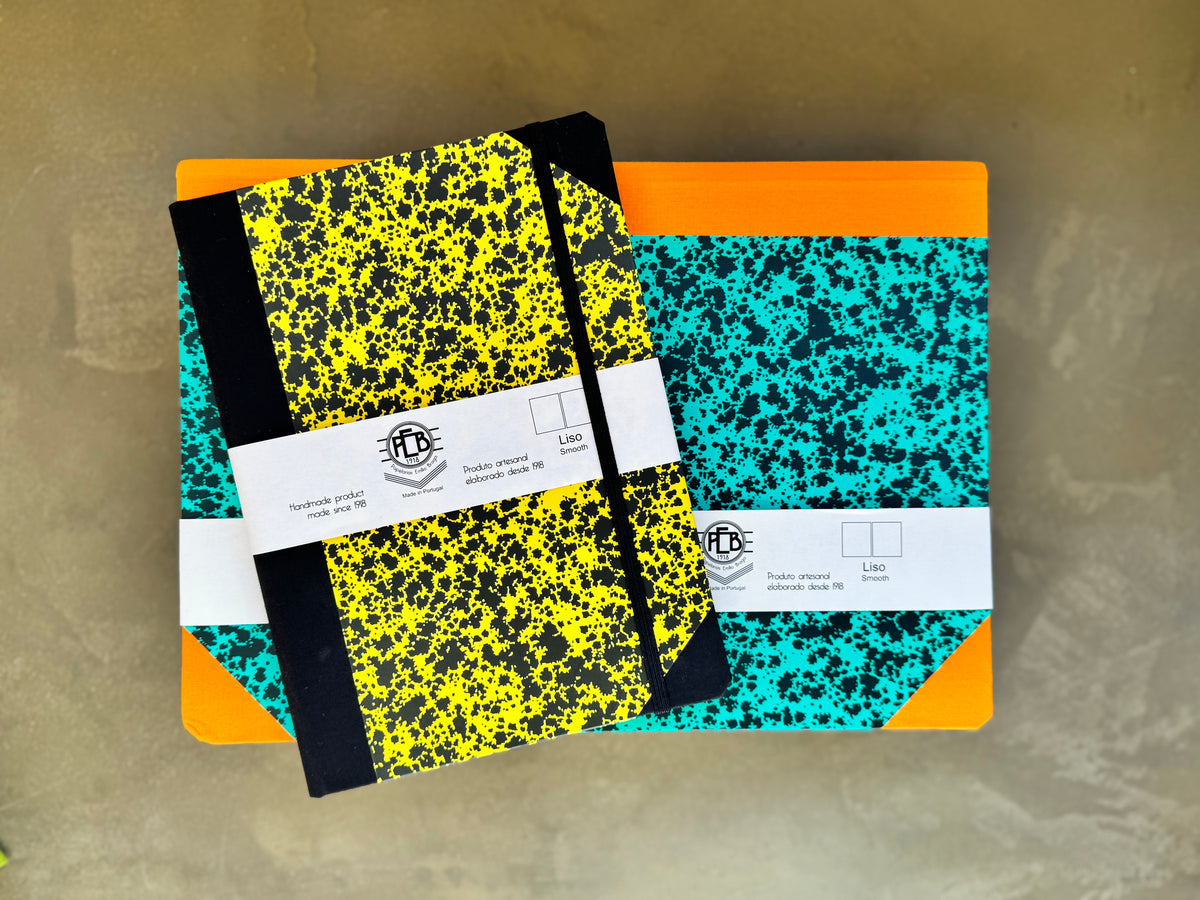 Two hardcover notebooks with black and yellow and black and turquoise/speckled covers, stacked on top of orange and white notebooks, with a white paper band across the middle. The notebooks are photographed from above on a textured beige surface.