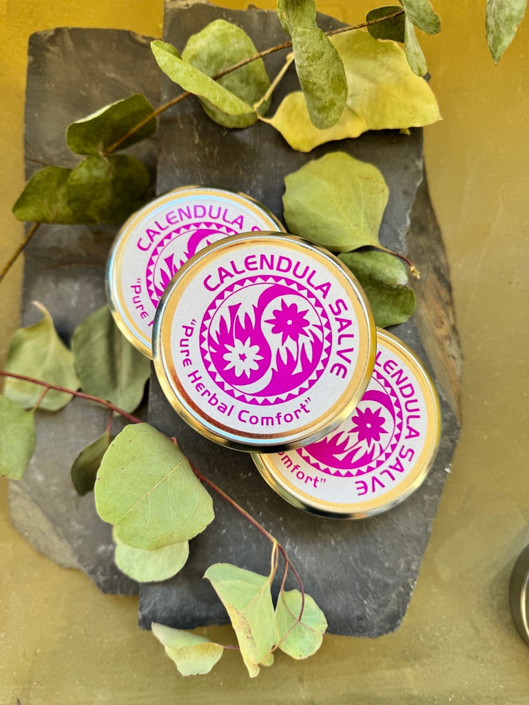 Three round metal tins of Calendula Salve arranged on a dark slate slab with green leaves, on a yellow surface. The tins have gold rims and a white label with a magenta/purple floral design and the text 'CALENDULA SALIVE' and 'Pure Herbal Comfort'.