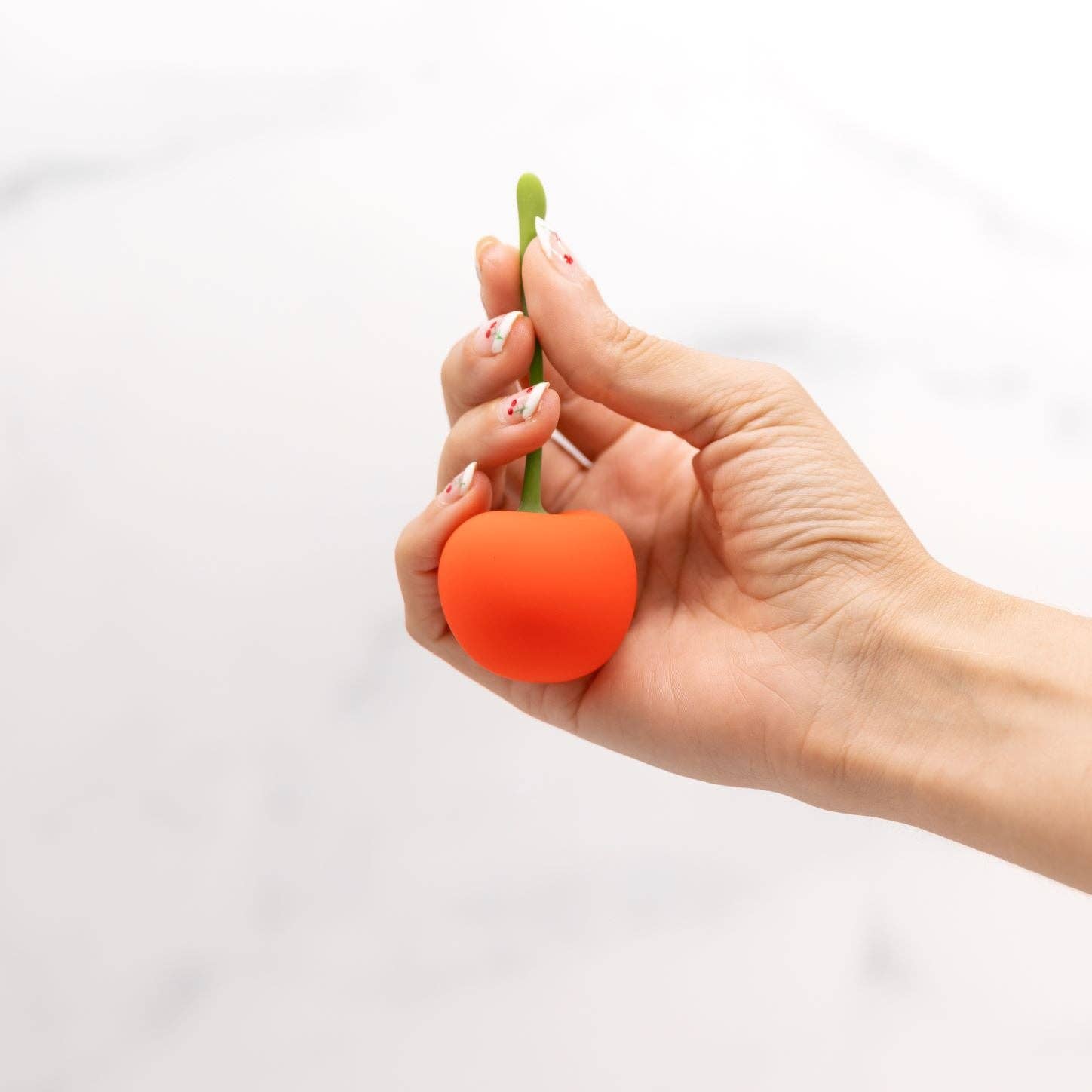 Hand holding a small cherry shaped vibrator with a green stem on a white background