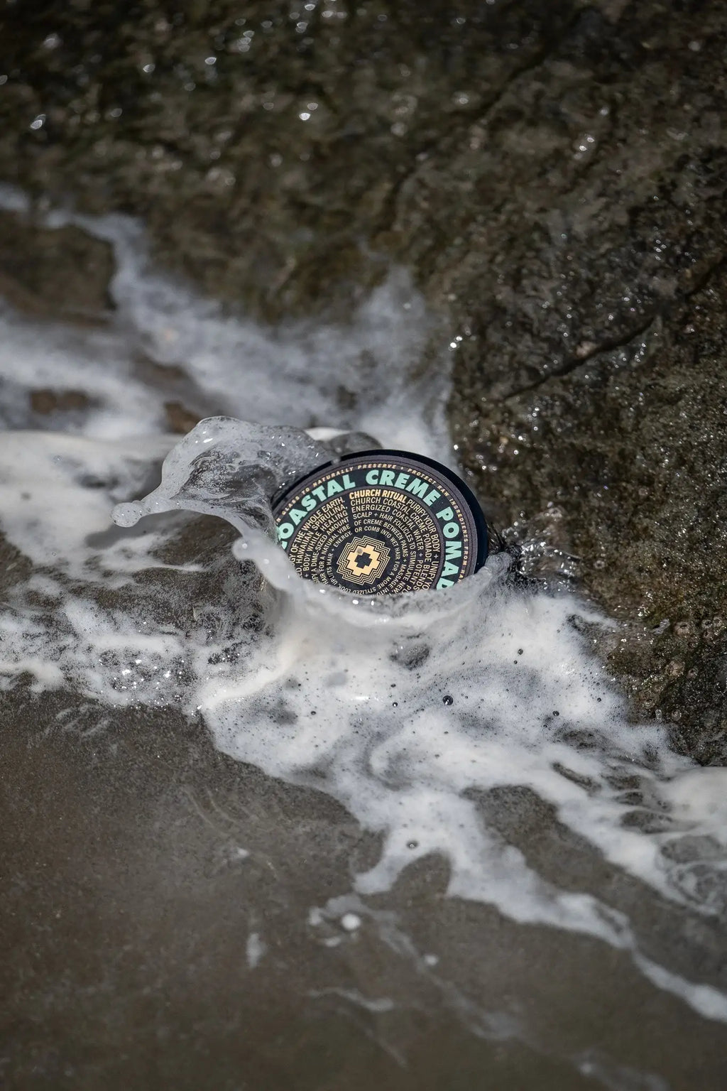 A circular black and white patterned patch of wax or adhesive floating in shallow surf with white foam and wet rocks around it. The patch has visible text reading 'COASTAL CREME POP.