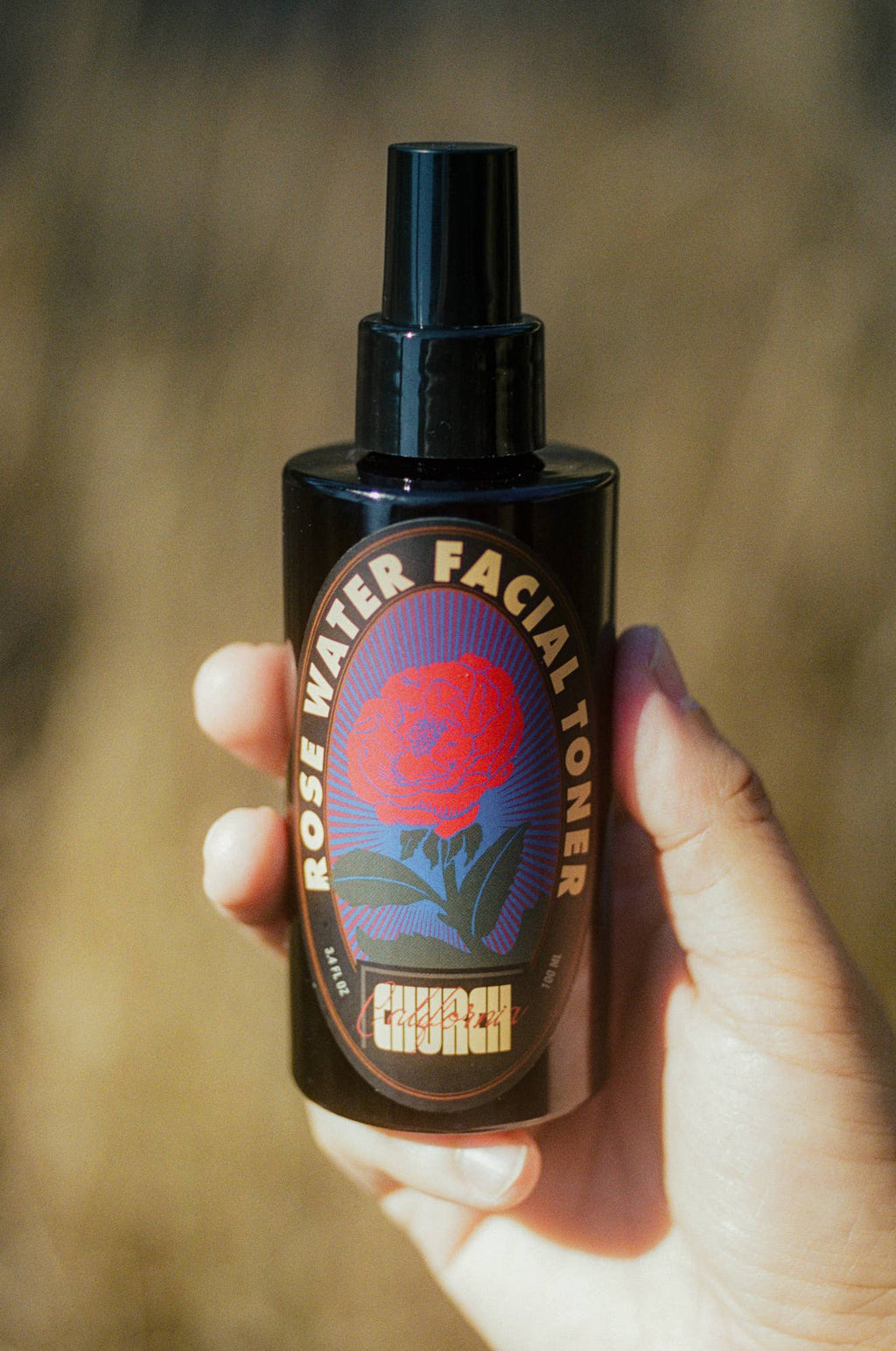 A hand holding a black spray bottle of Church Rose Water Facial Toner against a blurred warm brown background.