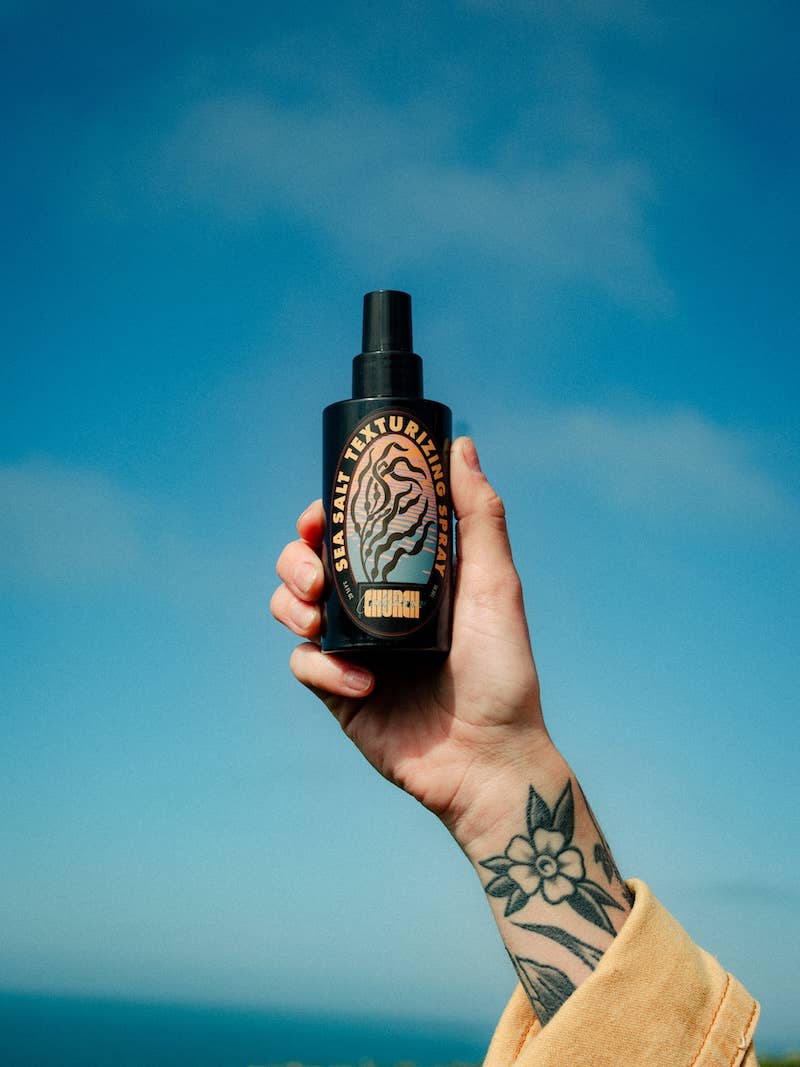 A hand with a floral tattoo holds up a black spray bottle of hair texture spray against a bright blue sky background.