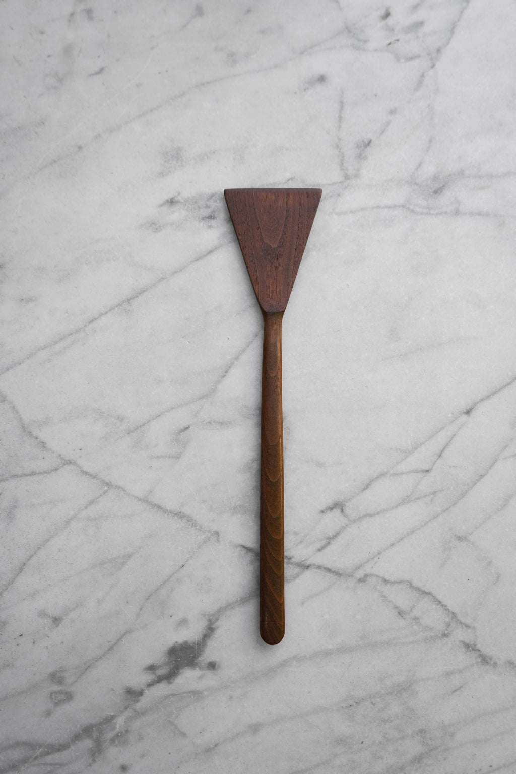 A dark brown wooden spatula with a triangular head and a long handle, placed vertically on a white marble surface with gray veining.