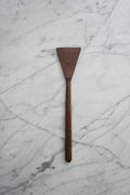 A dark brown wooden spatula with a triangular head and a long handle, placed vertically on a white marble surface with gray veining.