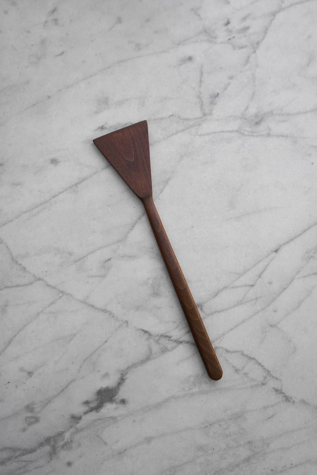 A dark brown wooden spatula with a triangular head and a long handle, placed diagonally on a white marble surface with gray veining.