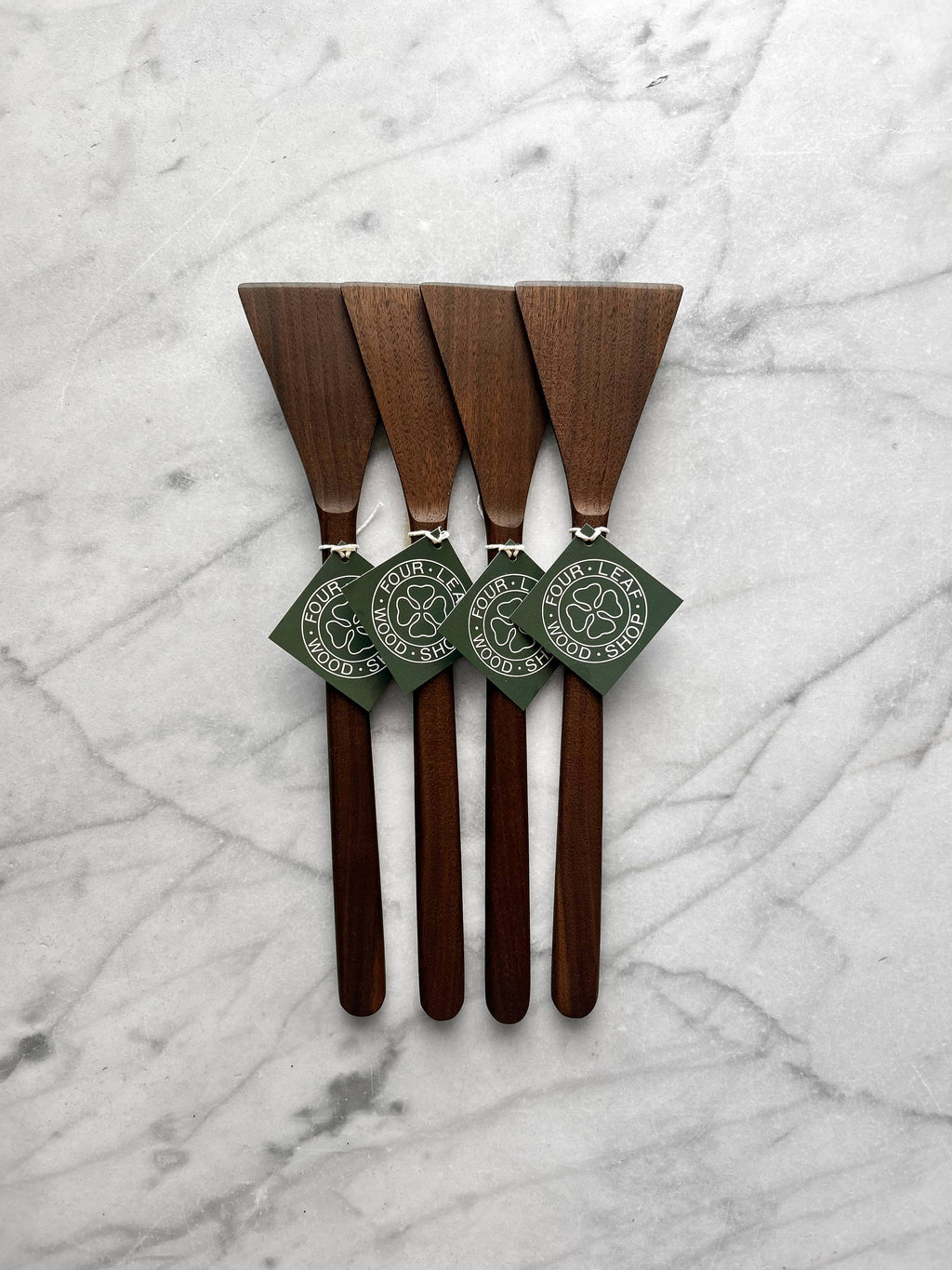 Four wooden serving spatulas with dark green paper tags, arranged vertically on a white and gray marble surface.