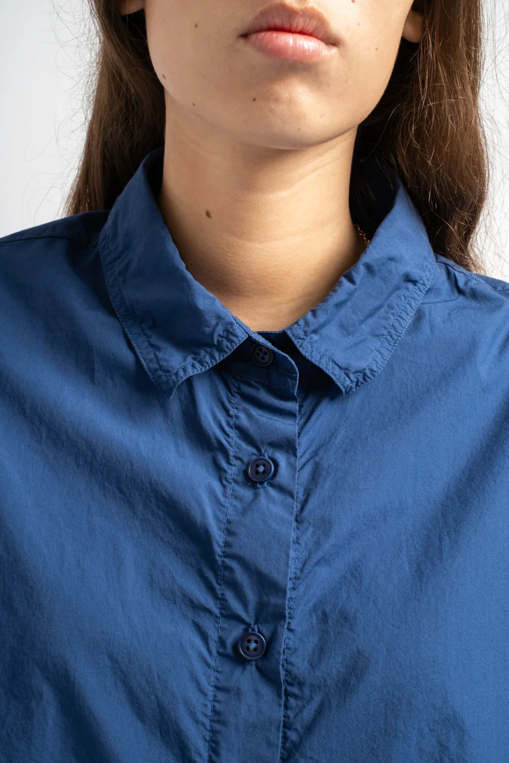 Close-up of a person wearing a blue button-front collared shirt, showing the collar, chest area, and part of the face against a light neutral background.