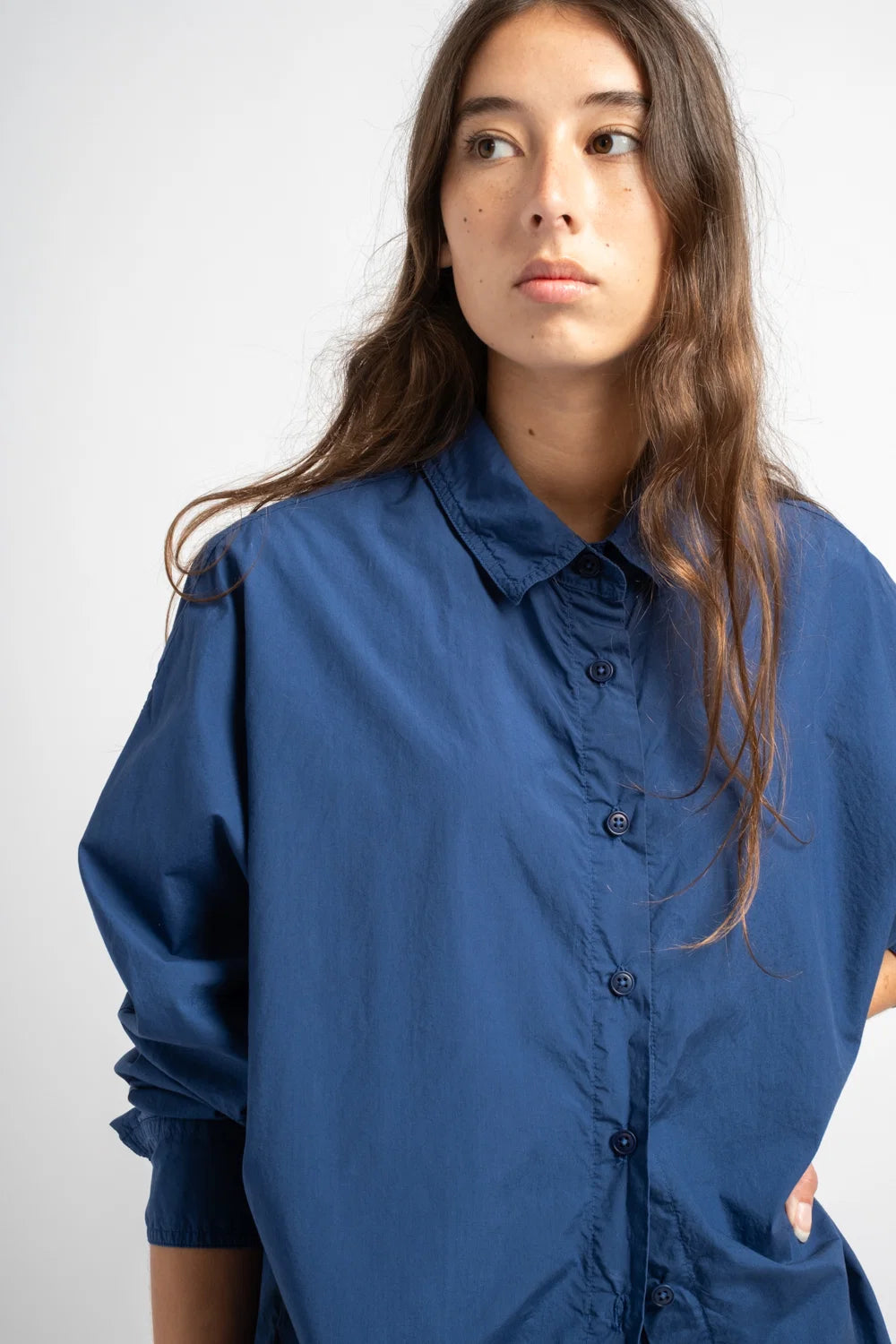 A woman wearing an oversized blue button-up shirt, photographed against a plain light gray studio background.