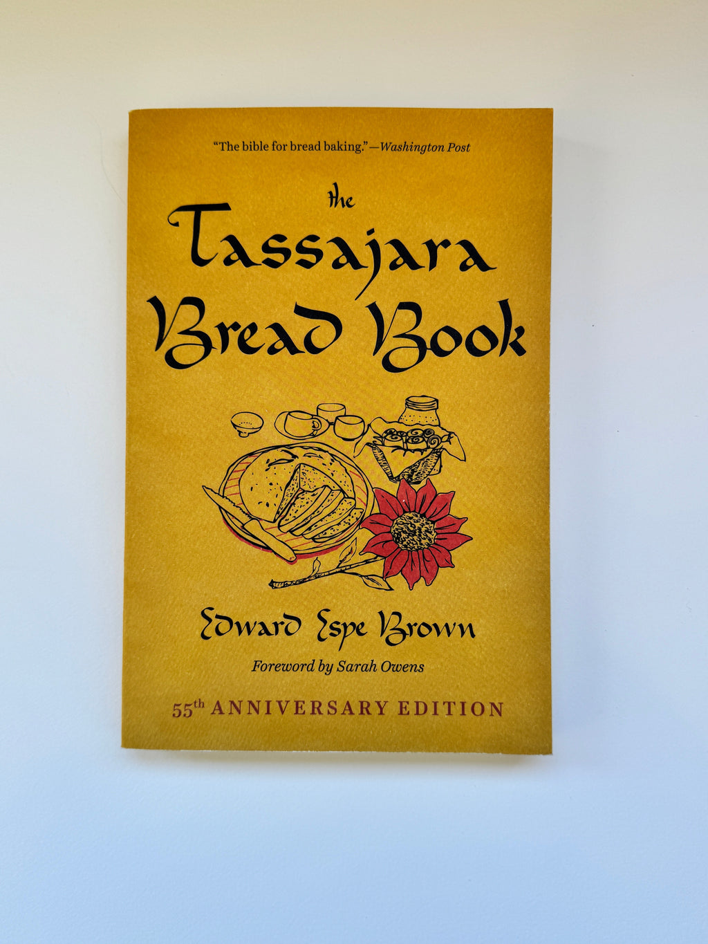 The Tassajara Bread Book 55th Anniversary Edition, featuring a mustard yellow cover with black text and illustrations of bread and a red flower, photographed against a light blue background.