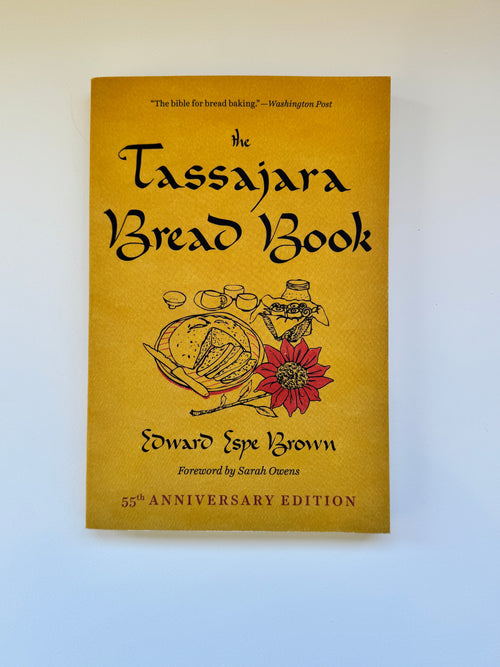 The Tassajara Bread Book 55th Anniversary Edition, featuring a mustard yellow cover with black text and illustrations of bread and a red flower, photographed against a light blue background.