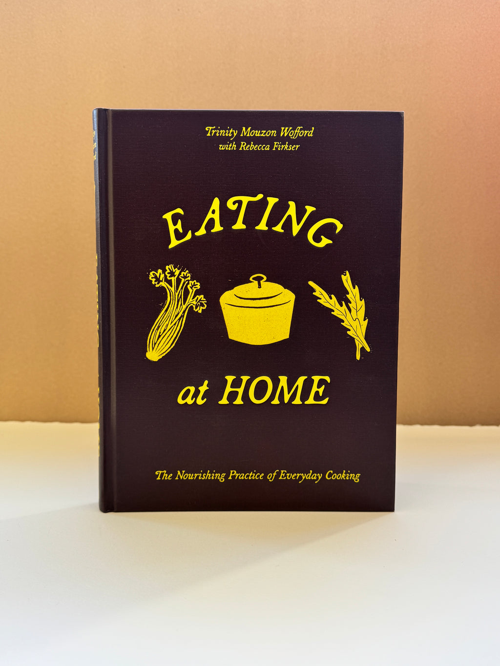 The front cover of the book 'Eating at Home' by Trinty Mason-Wifford and Rebecca Firthar, featuring a dark plum-brown background with yellow text and illustrations of food items, standing upright on a white surface against a beige background.